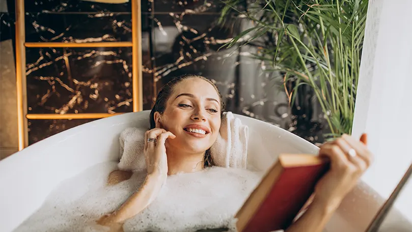 An image of a woman comfortably laying in a filled-up bathtub reading a book and enjoying herself